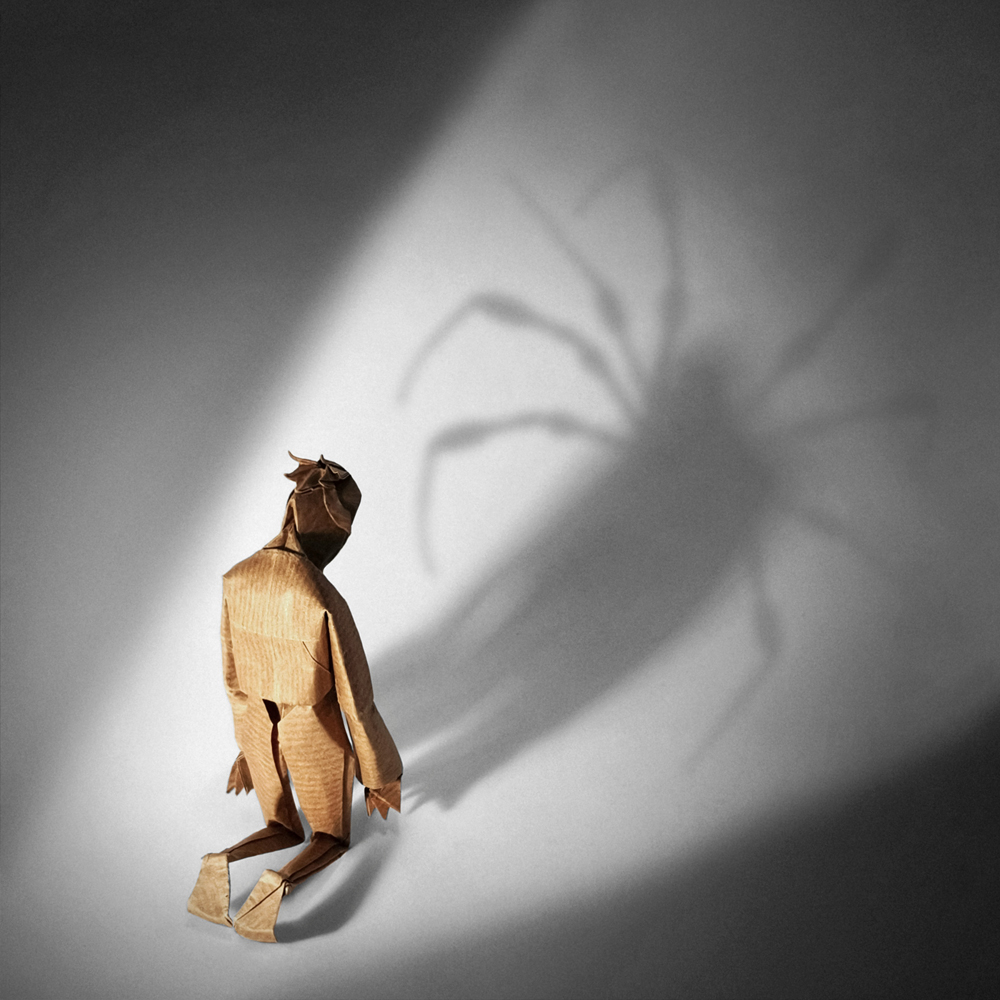 Origami of a little boy knelt over, head down, facing the shadow of a looming spider behind him.