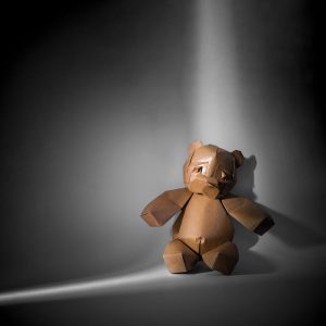 Origami of a teddy bear sitting in the back corner with a closet doors light crack shining on it.